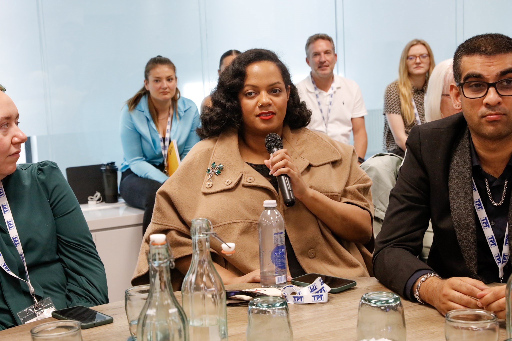 Laura and Ella, Worcestershire SLC members, pictured with Jagdeep, Birmingham and Black Country SLC member. They are seated at a table and Ella is talking into a microphone. Members of staff are pictured, seated, in the background.