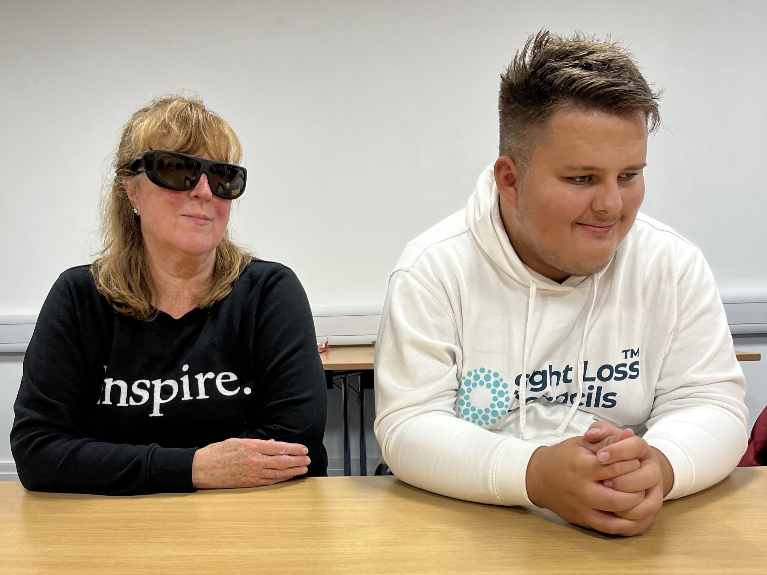 Lynne Rennison and Lloyd Sakr, pictured sitting next to each other during the launch meeting. Lynne is wearing sunglasses and looking at the camera smiling. Lloys is wearing his white SLC hoodies, and smiling whilst looking to the side.
