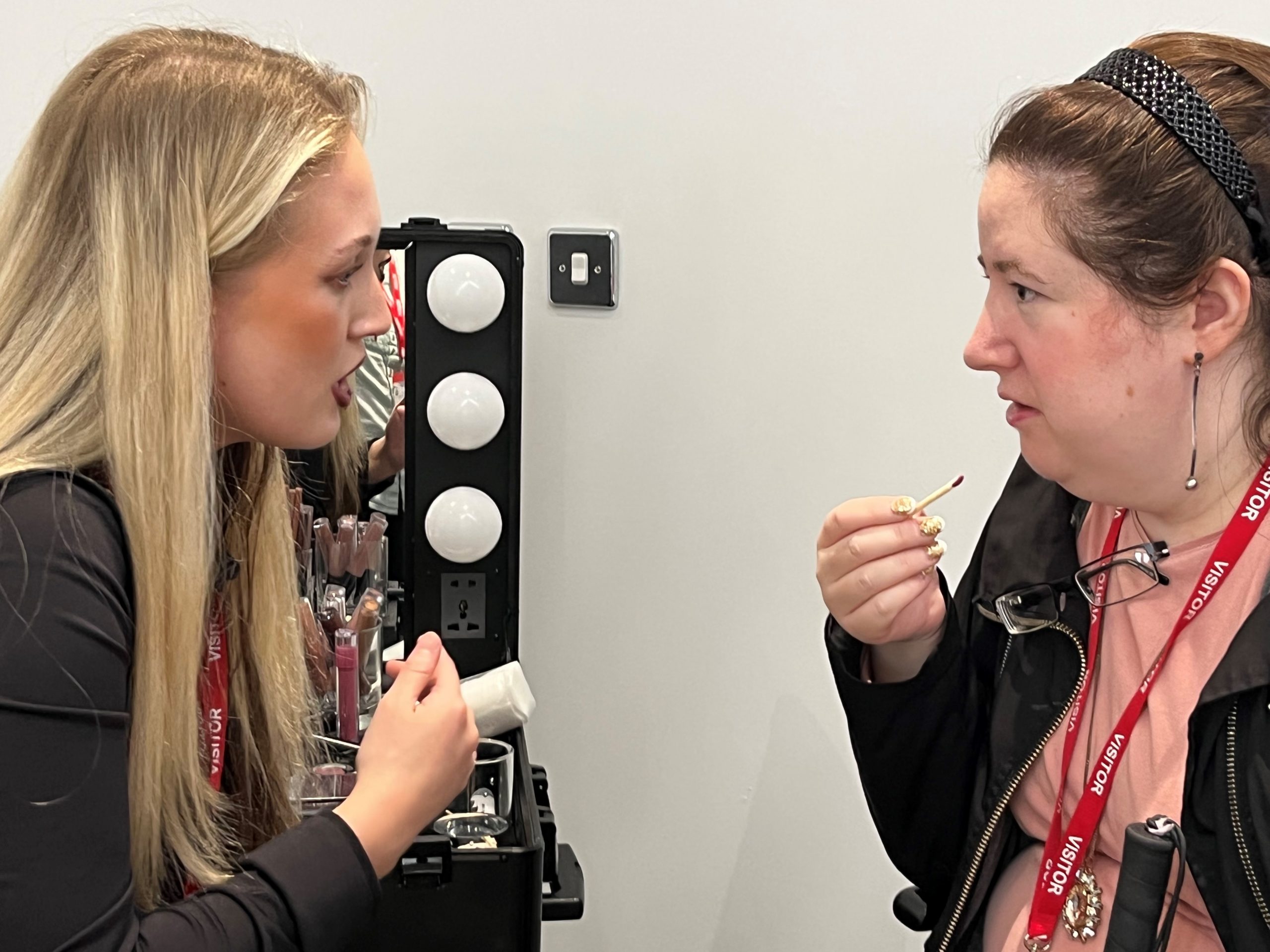 A makeup artist with attendee, Naomi Ullmann. Naomi is talking to the makeup artist, whilst holding a lip gloss brush up to her lips ahead of application.