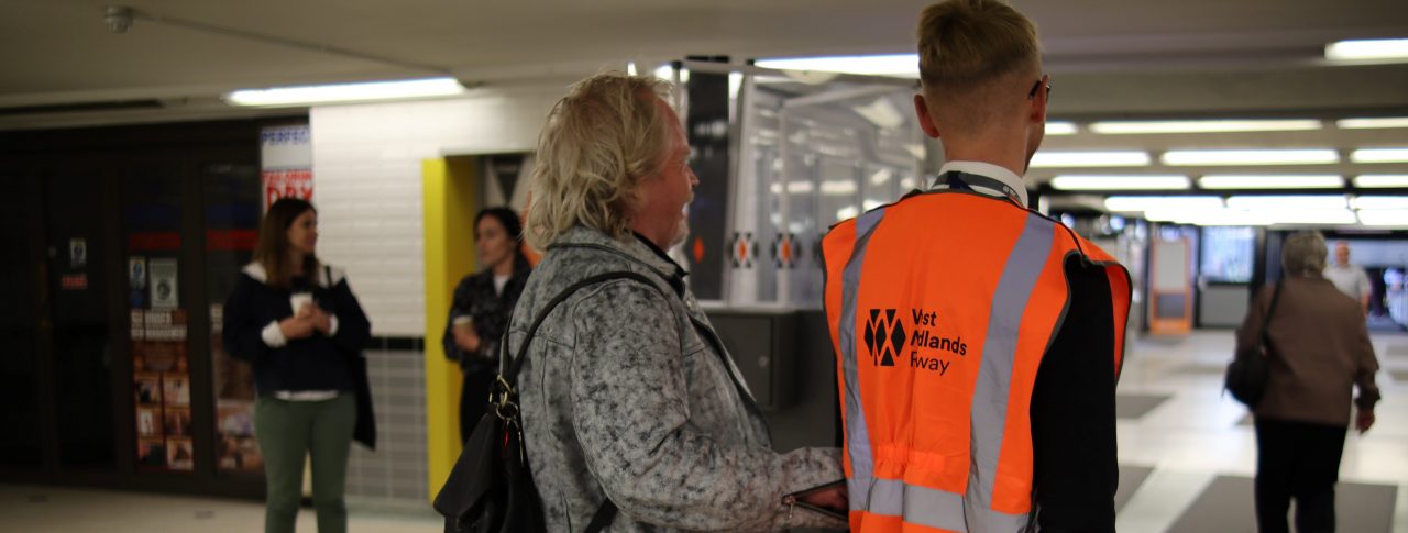 Steve Keith, Birmingham and Black Country SLC member, being guided through a ticket barrier by a member of staff. They have their backs to the camera.