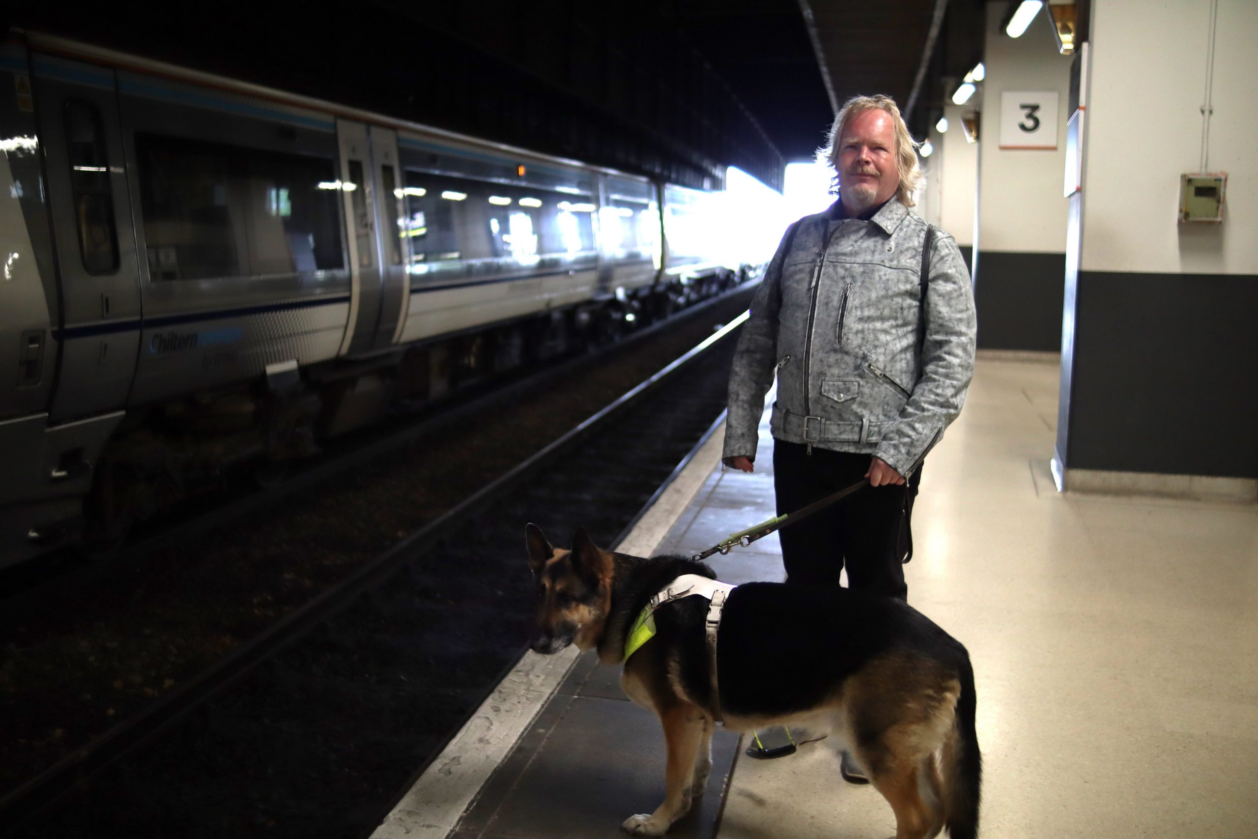 Steve Keith, Birmingham and Black Country Sight Loss Council member, standing on a train platform with his guide dog. Steve is looking at the camera, smiling. His guide dog is looking at the tracks. A train is shown on the opposite platform.