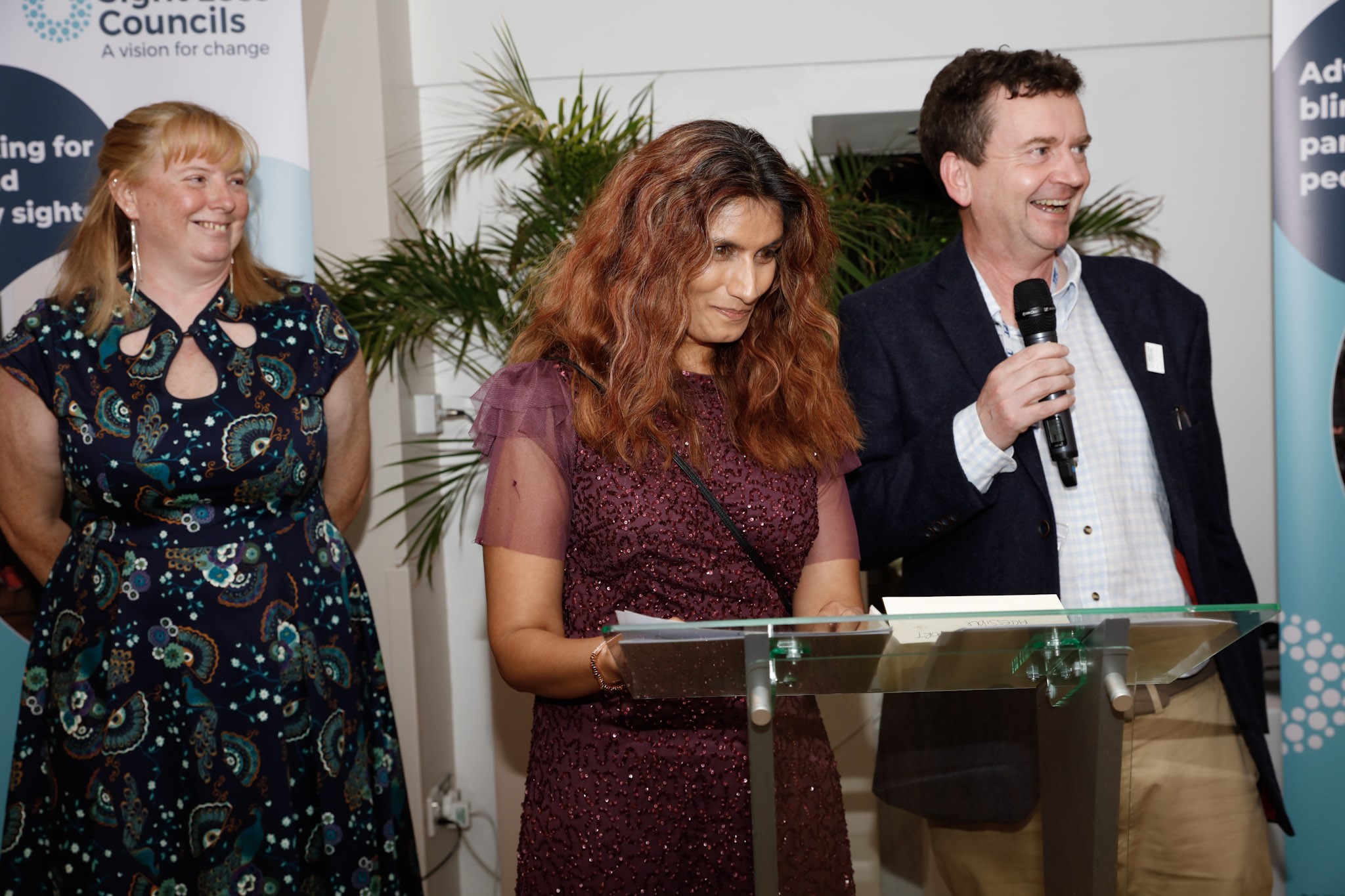 From left to right: Tricia Sail, Anela Wood, West of England SLC member, and Charles Colquhoun, CEO of Thomas Pocklington Trust. Anela is standing at a glass plinth, accepting the award. Charles is holding a microphone.