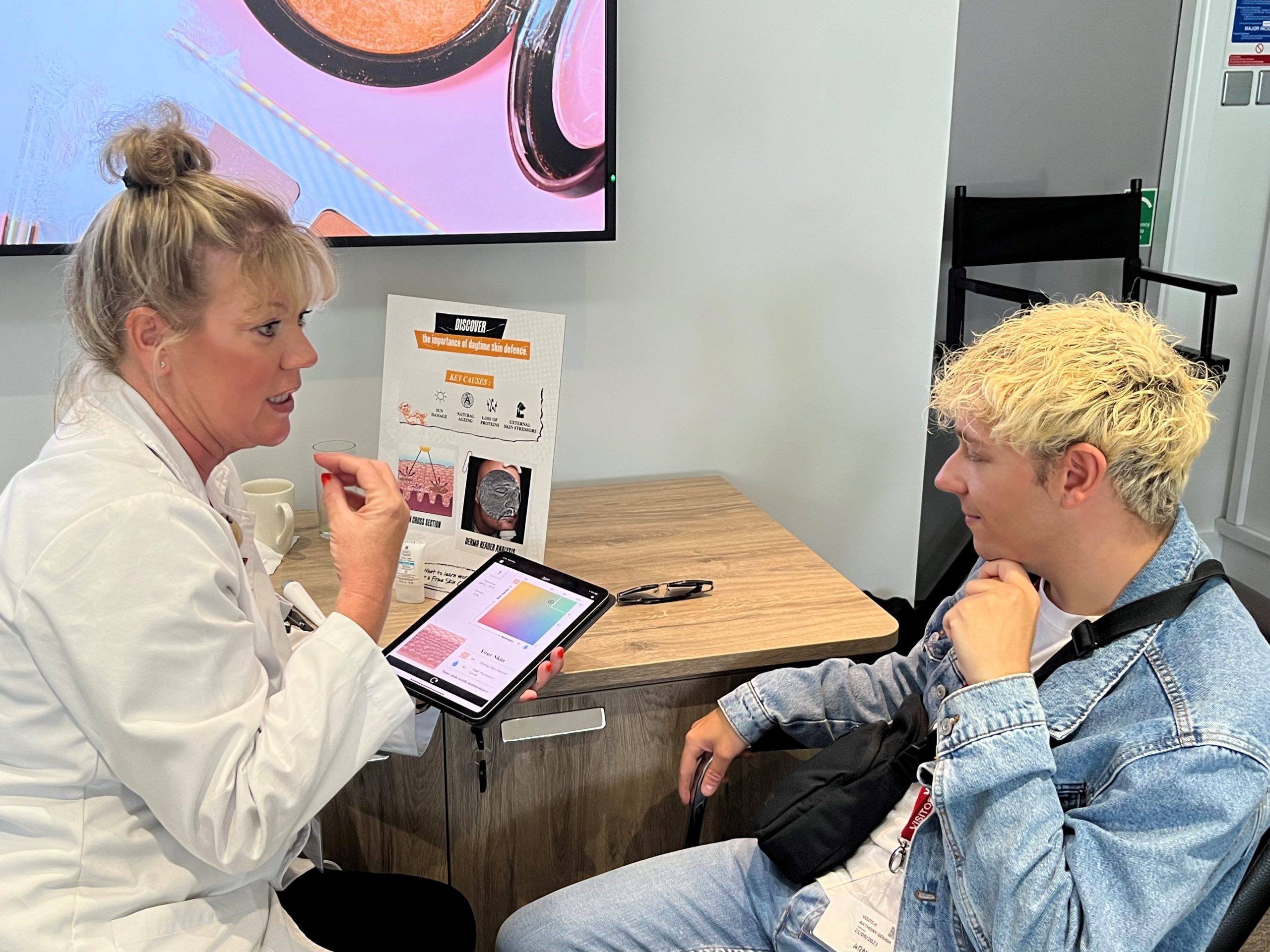 Anthony Gough, Greater Manchester SLC member, discussing skin care products during the make up event. They are seated at a desk, the technician is holding an iPad.