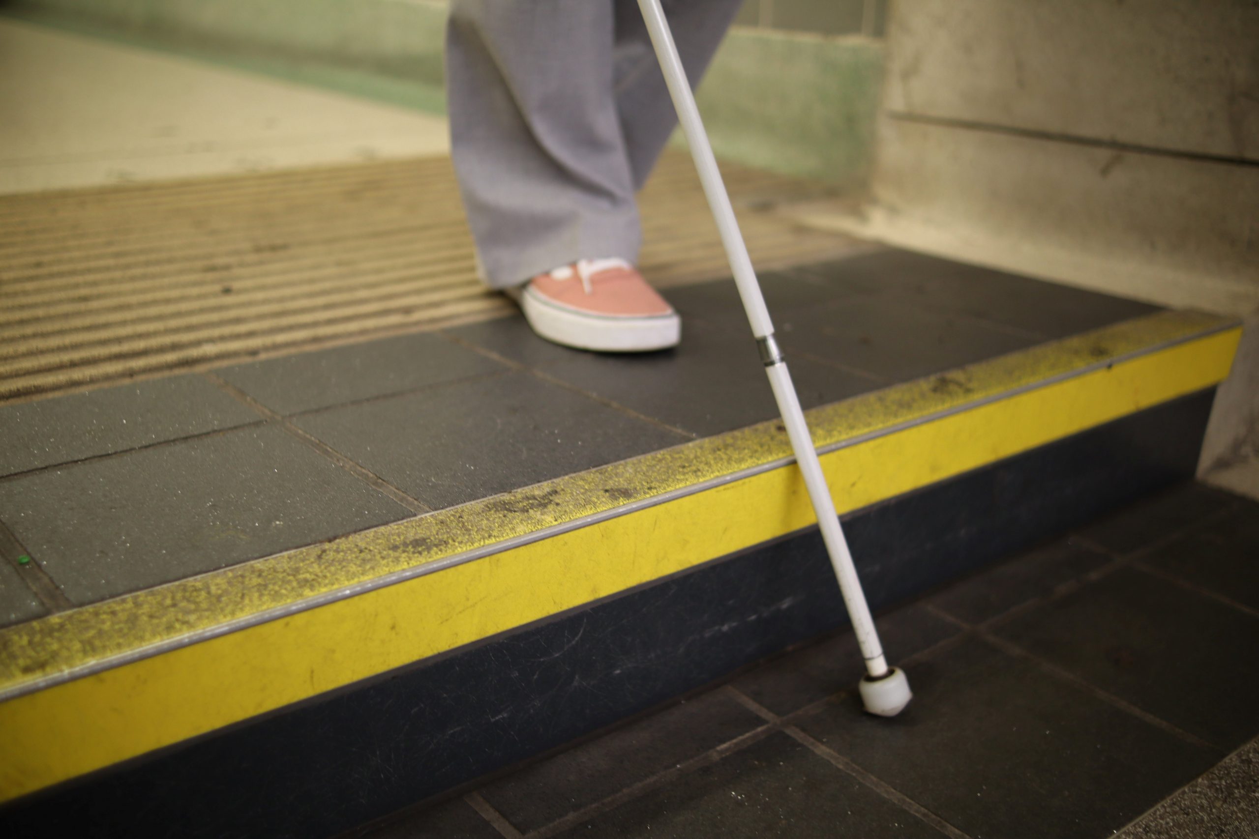 A lady's legs are shown from the shin down, on the top step. Her long cane is shown on the first step down as she prepares to step down.