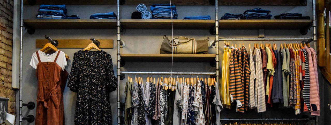 Clothes are hung across four clothes rails against a grey wall. Wooden shelves are above the rails, with denim clothing folded, and a canvas tote bag. The rails are industrial in style, to mimic old scaffolding poles.