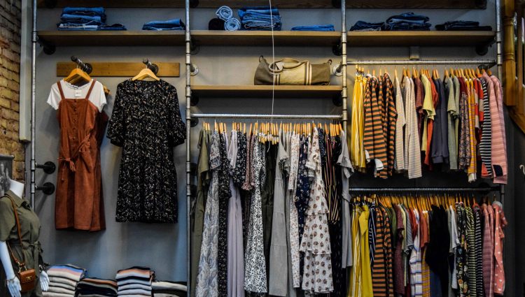 Clothes are hung across four clothes rails against a grey wall. Wooden shelves are above the rails, with denim clothing folded, and a canvas tote bag. The rails are industrial in style, to mimic old scaffolding poles.
