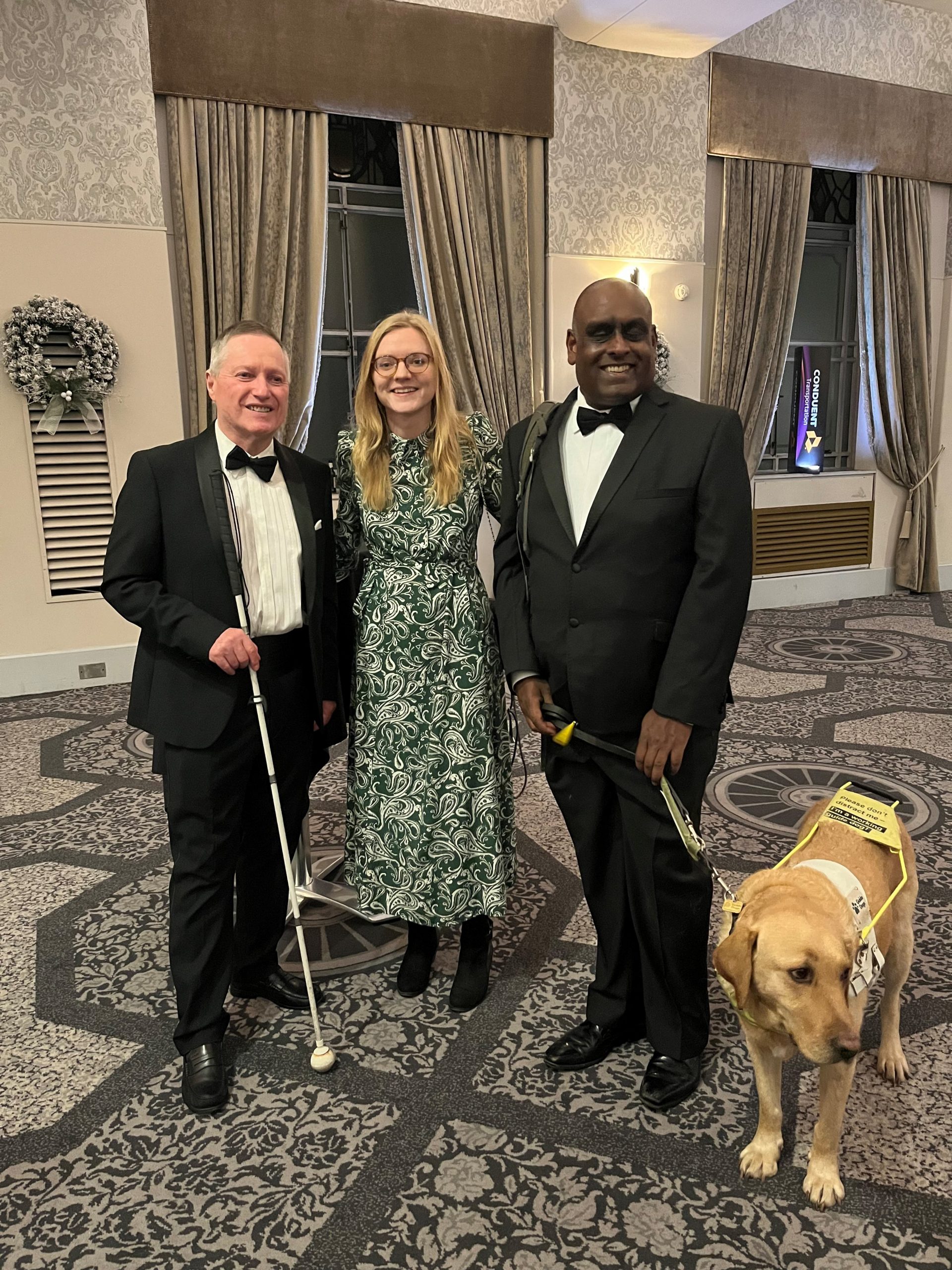 From left to right: Harry Meade, SW London SLC member, Lucy Williams, Senior Engagement Manager for South England, Haren Thillainathan, SW London SLC member, and Guide Dog Addi. They are stood in the foyer of the Connaught Rooms, London. Harry and Haren are wearing black tuxedos, and Lucy is wearing a full length, green and white dress. They are all facing the camera, smiling.