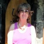 Photo of Karen sitting on a wooden chair in the sun. She is wearing sunglasses and a summer top, smiling at the camera.