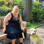 Leanne, London SLC member is sitting on a park bench with her guide dog. She is looking forwards, laughing.