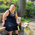 Leanne, London SLC member is sitting on a park bench with her guide dog. She is looking forwards, laughing.