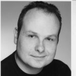 Black and white headshot of Liam O'Carroll. His head is turned slightly to the side and he is smiling.