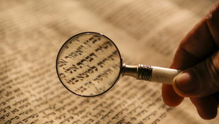 A male’s hand, holding a magnifying glass over an ancient document, written in script.