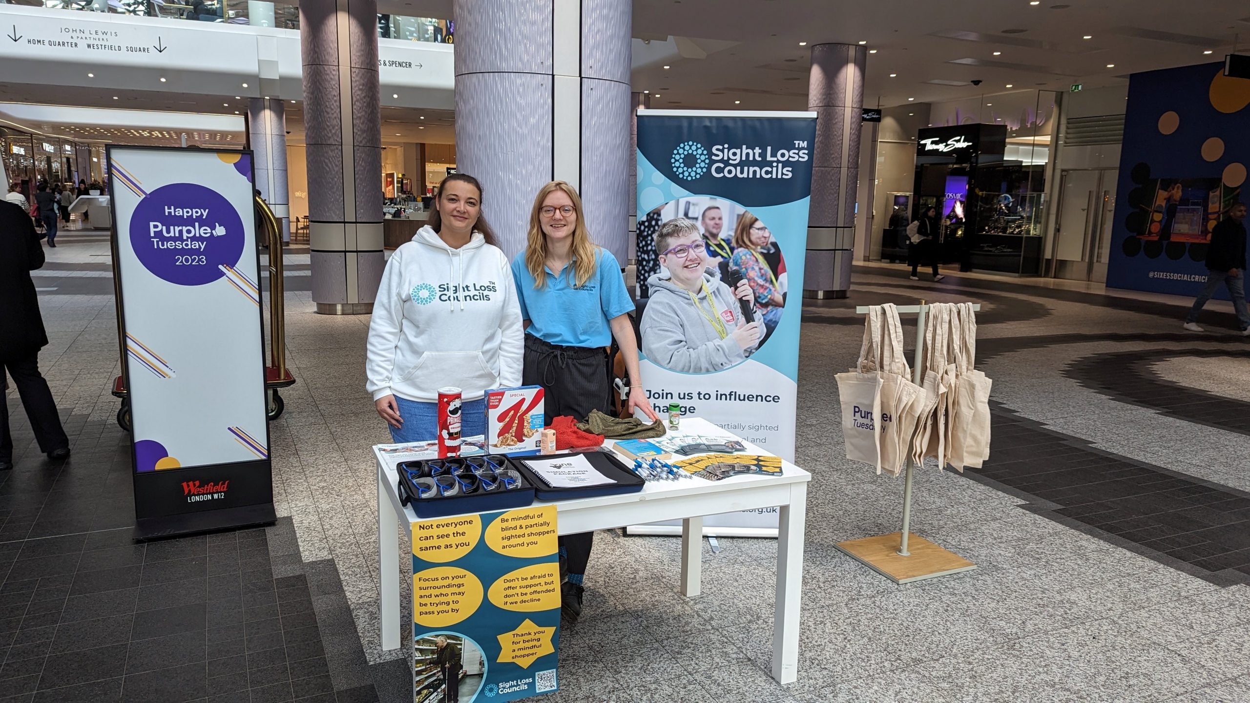 Nikki Hughes, SW London SLC member, and Lucy WIlliams, Senior Engagement Manager for South England, stood at their table in Westfield London. To the left is a Purple Tuesday 2023 banner, and to the right is the Sight Loss Councils banner. On the table are various products, a case with sim specs in it, and the 'Mindful shopper' poster.