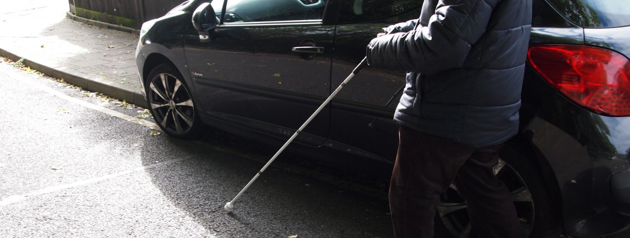 A blind man using a white cane, is walking on the road, around a car which is parked on the pavement.