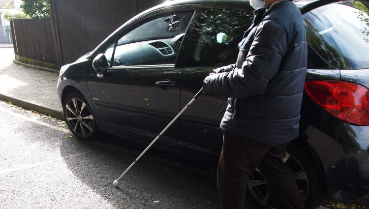 A blind man using a white cane, is walking on the road, around a car which is parked on the pavement.