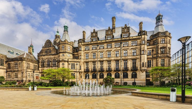 Landscape image of Sheffield Town Hall, a Grade 1 listed building built with Stoke stone and ornate carvings. In front of the town hall is a circular fountain, grass and trees.
