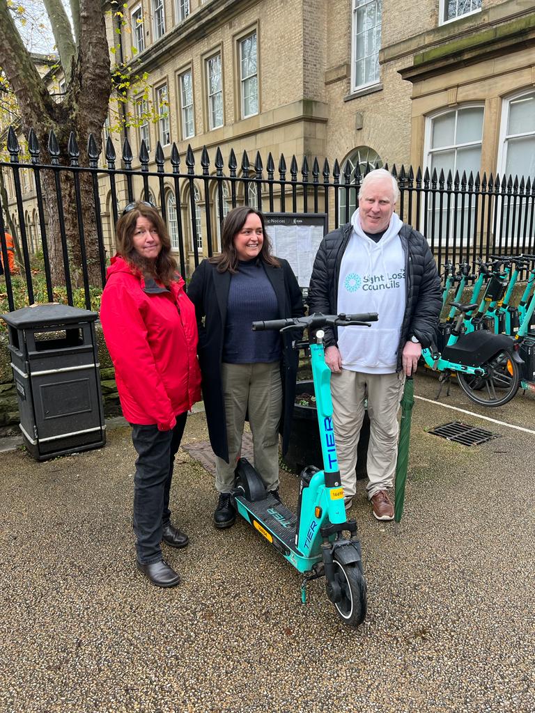 SLC volunteer Hilary Boon is stood with Jessica Hall, Head of Public Policy, UK, TIER, and Iain Mitchell, Senior Engagement manager for North England. They are standing next to an e-scooter to the side of a row of e-scooters in a parking bay.