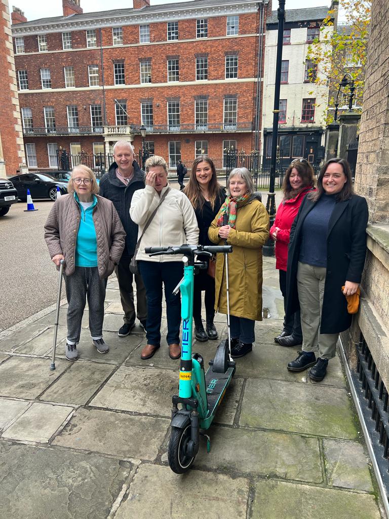 From left to right: Denise Peat, Iain, SLC member Verity Peat, Belle Whitely, SLC Coordinator for Yorkshire and Humberside, SLC members Josie Clarke and Hilary Boon, and Jessica Murphy, Head of Public Policy, UK, TIER. They are all standing behind an e-scooter following the AVAS trial in York.