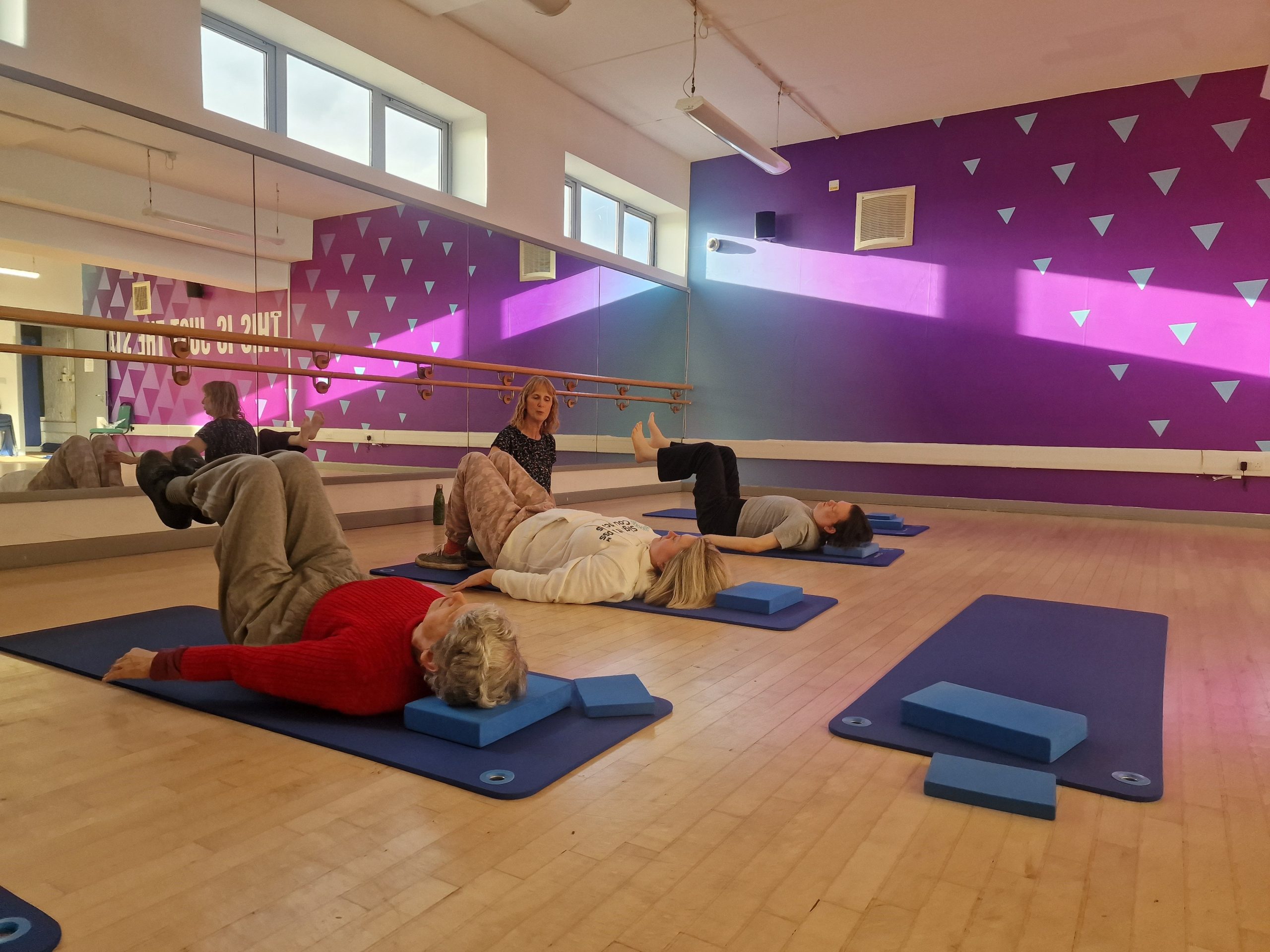 Three of our Let's Get Active attendees during the VI Yoga session. They are laying down, with their knees bent in the air. The instructor is knelt in front of them. Sunlight is flooding the studio.