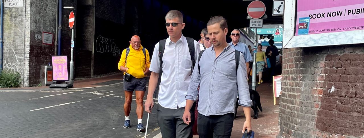 Dave Smith, Engagement Manager for South East England, is shown walking on a pavement in Brighton with his long cane. His support worker is walking next to him, and several people are walking on the path behind him.