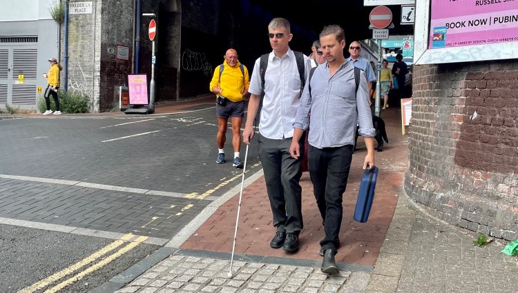 Dave Smith, Engagement Manager for South East England, is shown walking on a pavement in Brighton with his long cane. His support worker is walking next to him, and several people are walking on the path behind him.