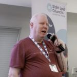 Iain Mitchell, Senior Engagement Manager for the North, speaking into a microphone. A SLC banner is behind him.