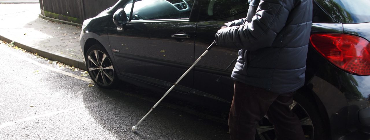 A blind man using a white cane, is walking on the road, around a car which is parked on the pavement.