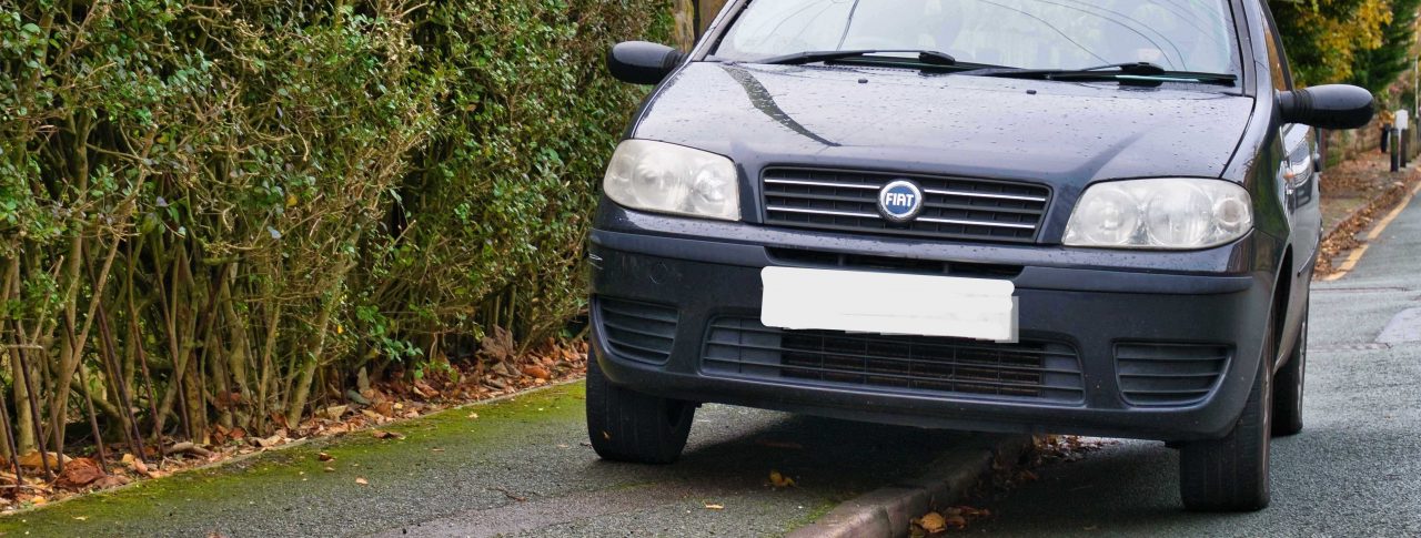 A black car which has parked on a pavement next to a tall hedge, taking up over half of the pavement.