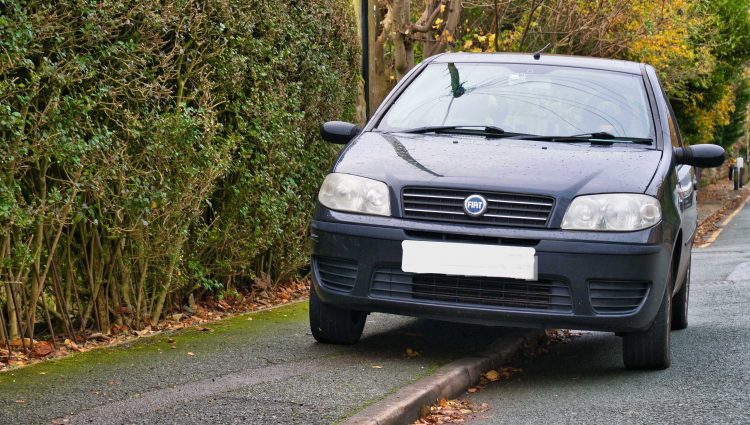 A black car which has parked on a pavement next to a tall hedge, taking up over half of the pavement.