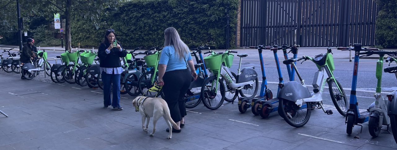 Leanne Best, London SLC member, is standing next to a bank of Lime e-bikes and e-scooters with her back to the camera. She is facing a female camera operator and walking towards the camera with her guide dog.