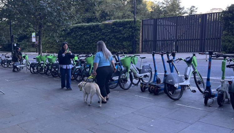 Leanne Best, London SLC member, is standing next to a bank of Lime e-bikes and e-scooters with her back to the camera. She is facing a female camera operator and walking towards the camera with her guide dog.