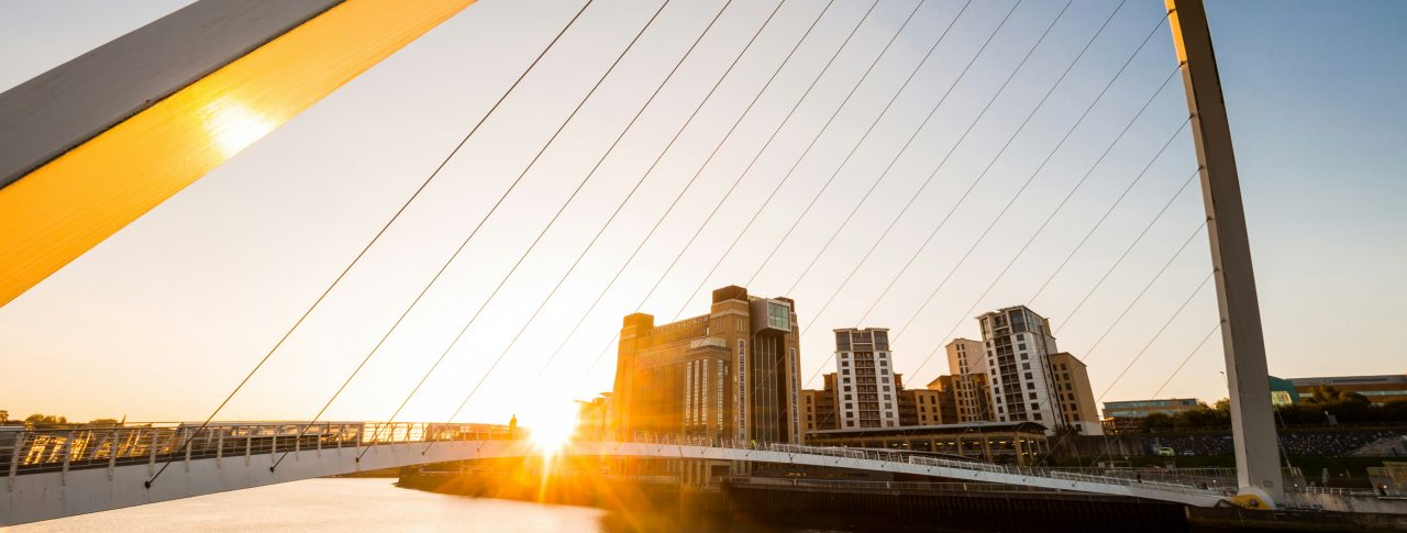 A close up of The Tyne Bridge in Newcastle, taken at sunset. Golden sunlight is flooding the bridge. A group of tall buildings can be seen in the distance.