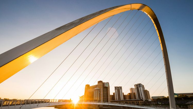 A close up of The Tyne Bridge in Newcastle, taken at sunset. Golden sunlight is flooding the bridge. A group of tall buildings can be seen in the distance.