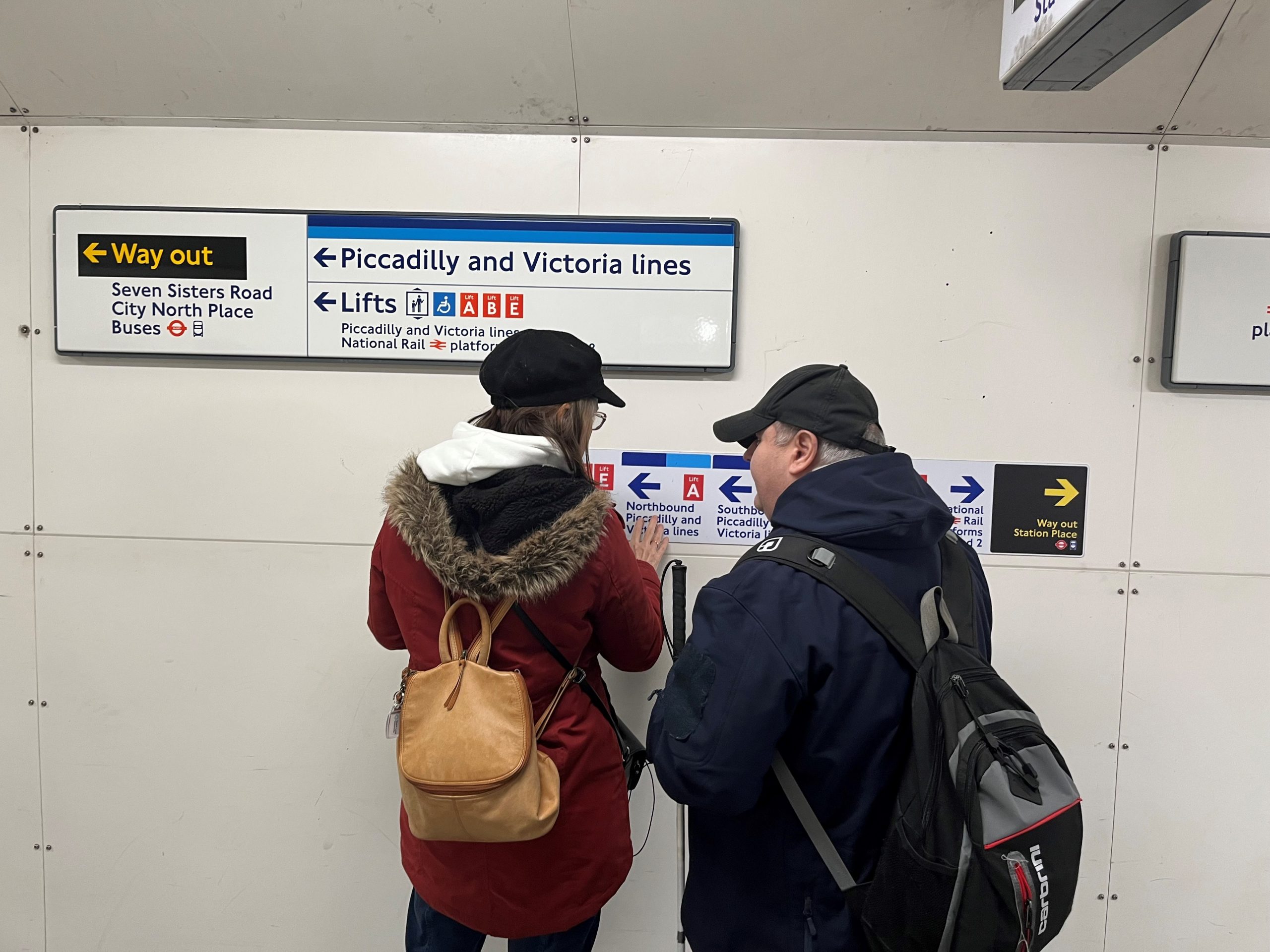 Vicky and Emmanuel, SLC members, are stood with their backs to the camera, whilst reading a sign to the underground. Vicky is touching the sign with her fingers as she reads it.
