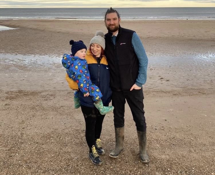 Jack photographed on a beach with his wife Becky and son Finlay. They are all wrapped up warm, Jack is smiling at the camera and Becky is looking down at Finlay - who is sitting on her hip.
