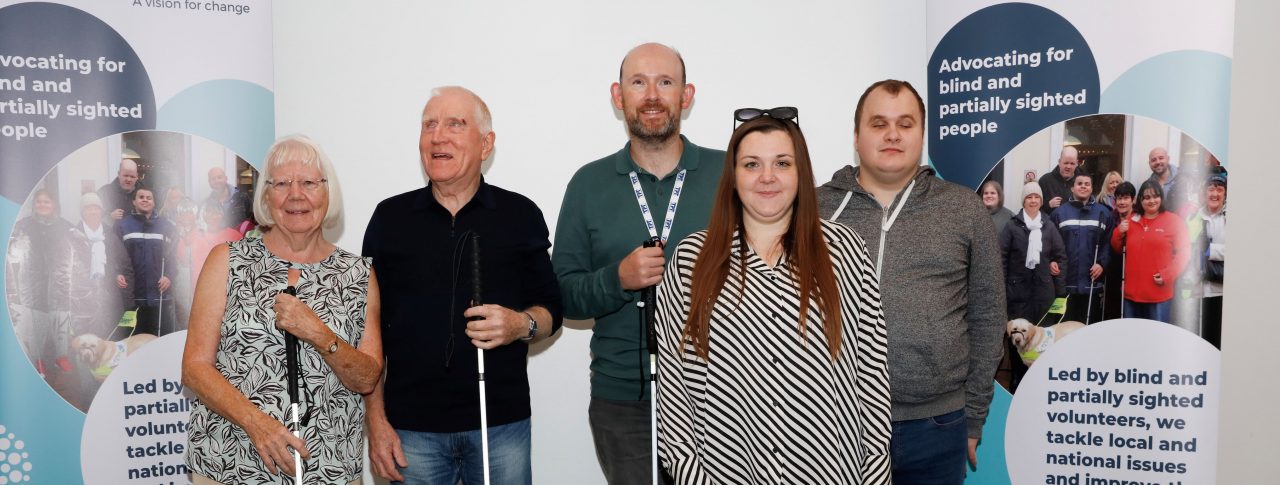 From left to right: SLC members Eileen, Phil, Stefan, and Hubert, pictured with Sam and guide dog Lizzie at the 2023 SLC Conference. They are stood together next to our SLC banners, all smiling at the camera.