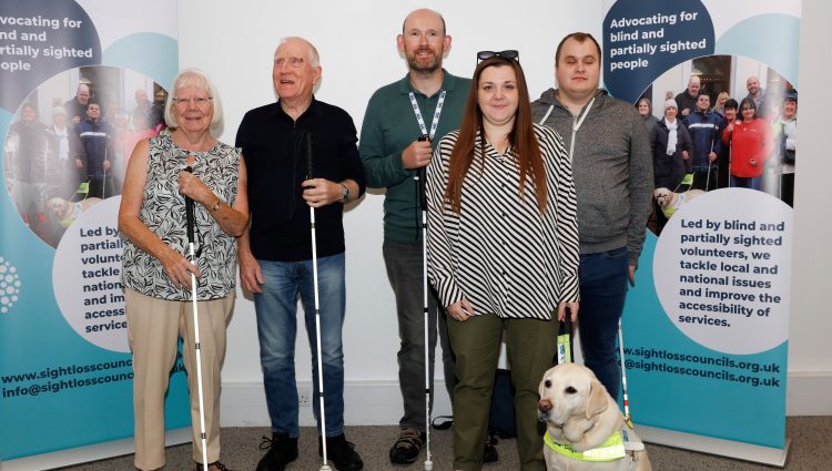 From left to right: SLC members Eileen, Phil, Stefan, and Hubert, pictured with Sam and guide dog Lizzie at the 2023 SLC Conference. They are stood together next to our SLC banners, all smiling at the camera.