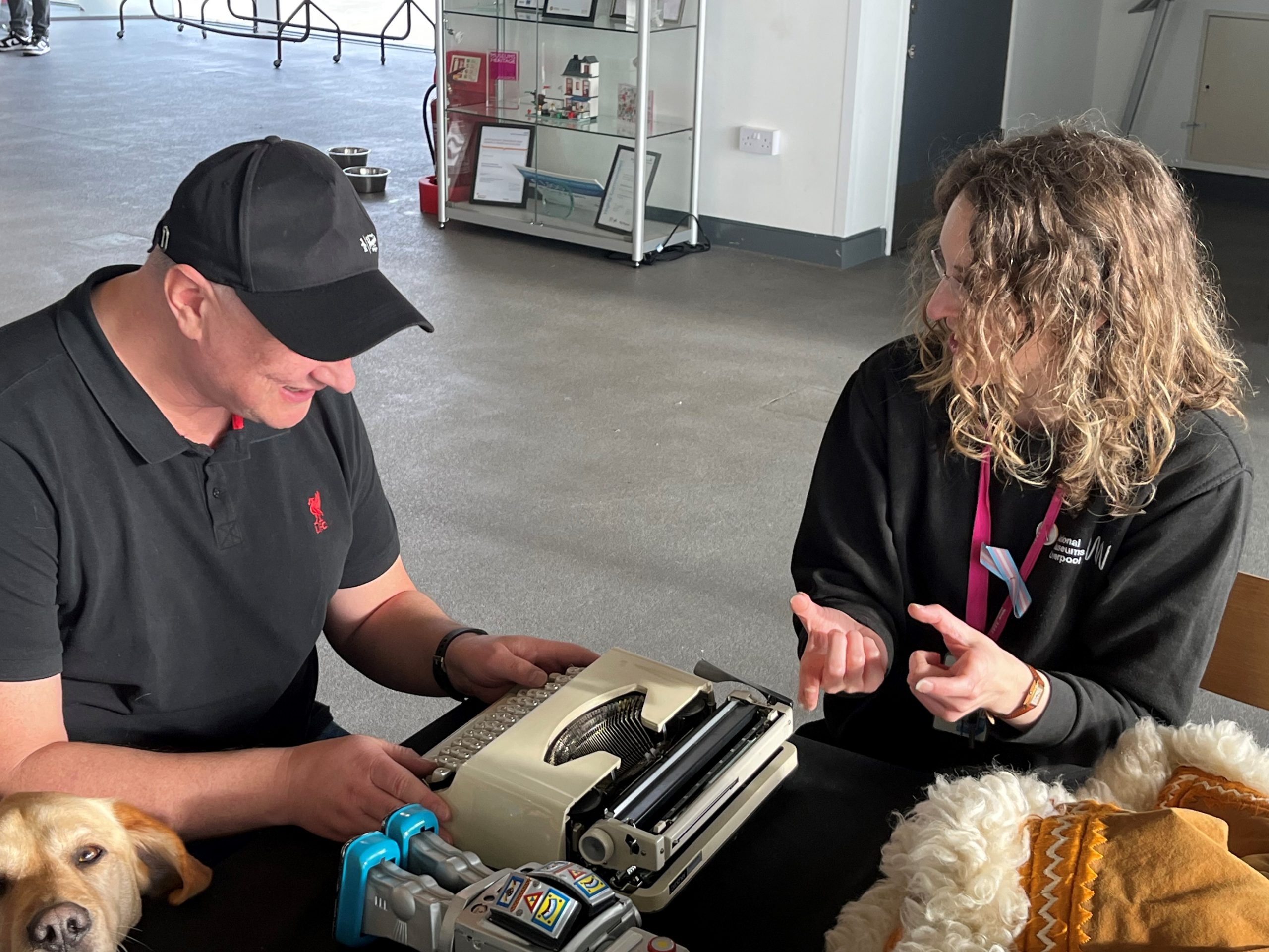 A delegate at Merseyside SLC's accessible museum event during the touch-tour element of the event. He is holding a typewriter. Other props can be seen on the table, and his guide dog's head is poking in the corner of the image.