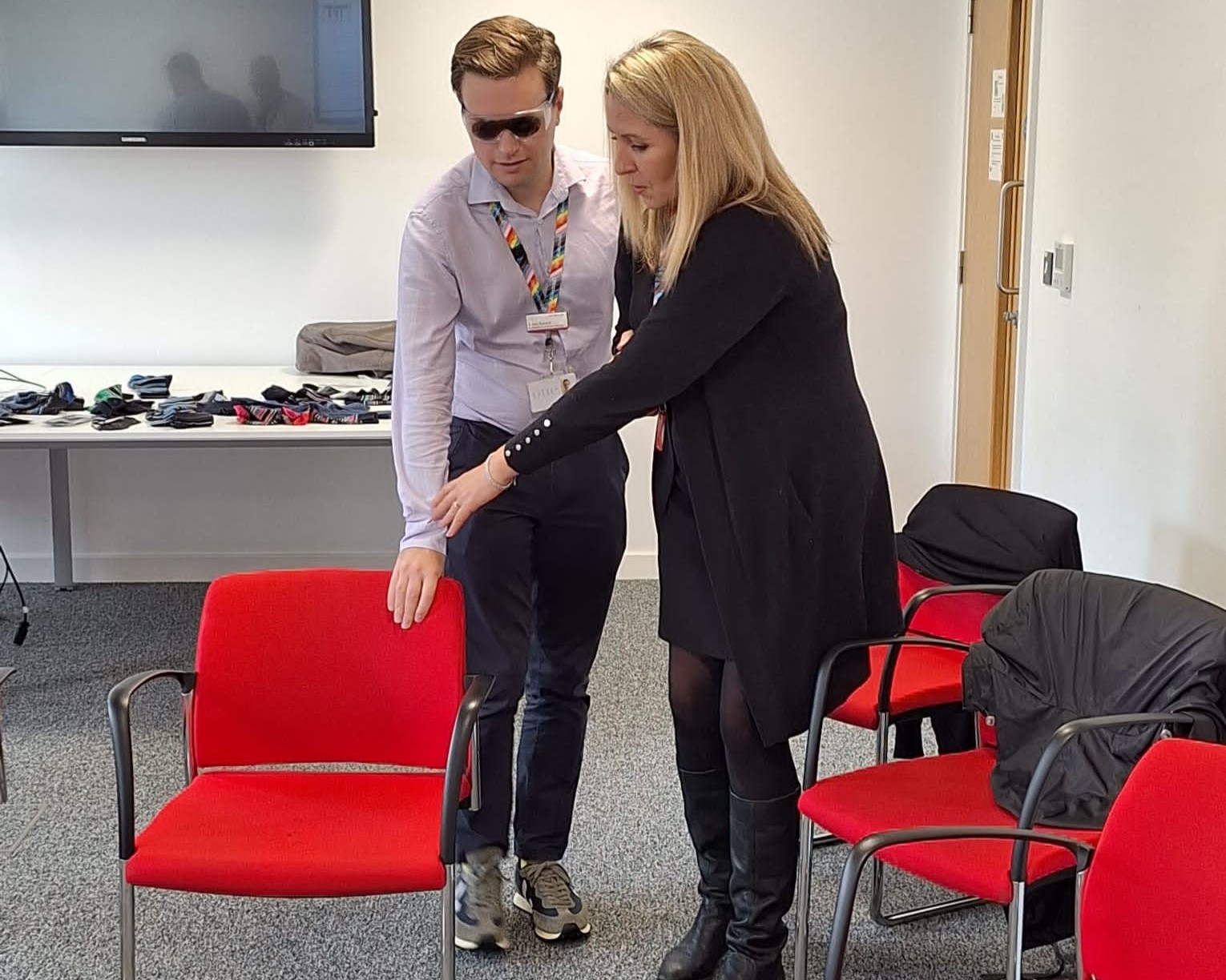 Greater Anglia staff during the sighted guide session. A male staff member is wearing sim-specs, and being guided to a red chair by a female colleague.
