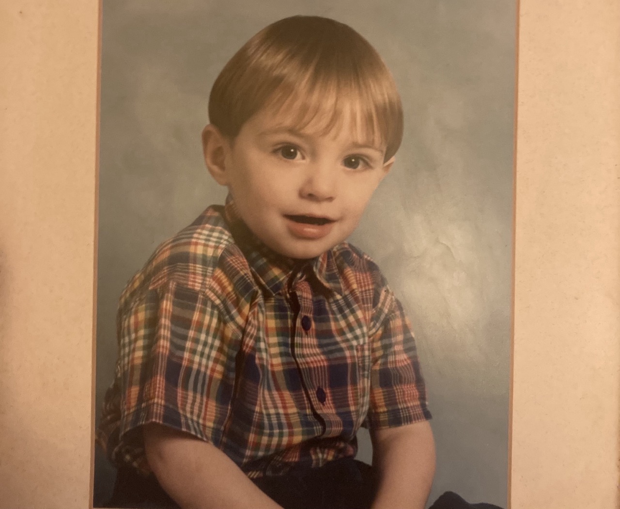 A photo of Jack, aged three years old. He is siting down, wearing a short sleeve plaid shirt. He is smiling at the camera.