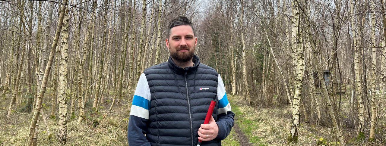Jack Moffat, SLC Engagement Manager for North East England. He is standing in a woods, surrounded by trees. Jack is holding his long cane and smiling at the camera.