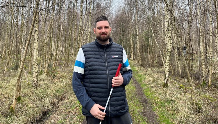 Jack Moffat, SLC Engagement Manager for North East England. He is standing in a woods, surrounded by trees. Jack is holding his long cane and smiling at the camera.