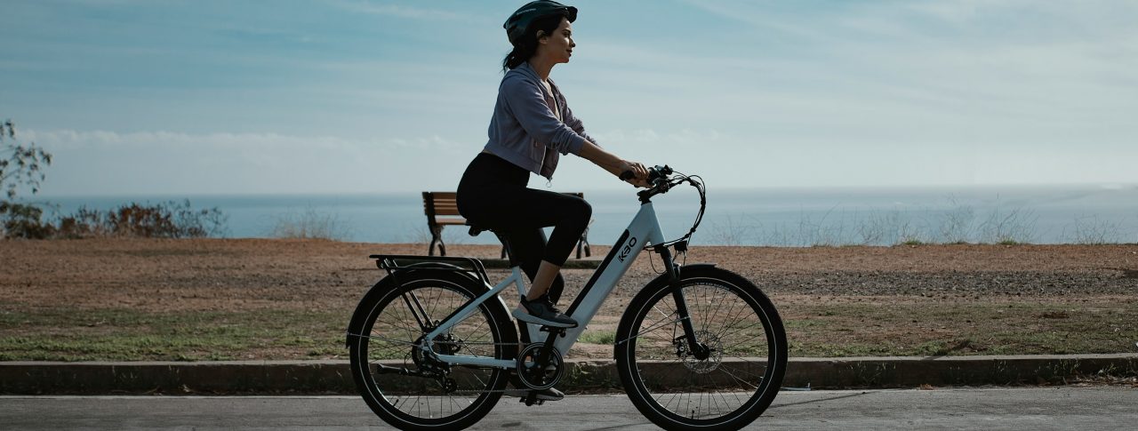 A lady riding a white e-bike on a sunny day, with the sea glistening in the distance.