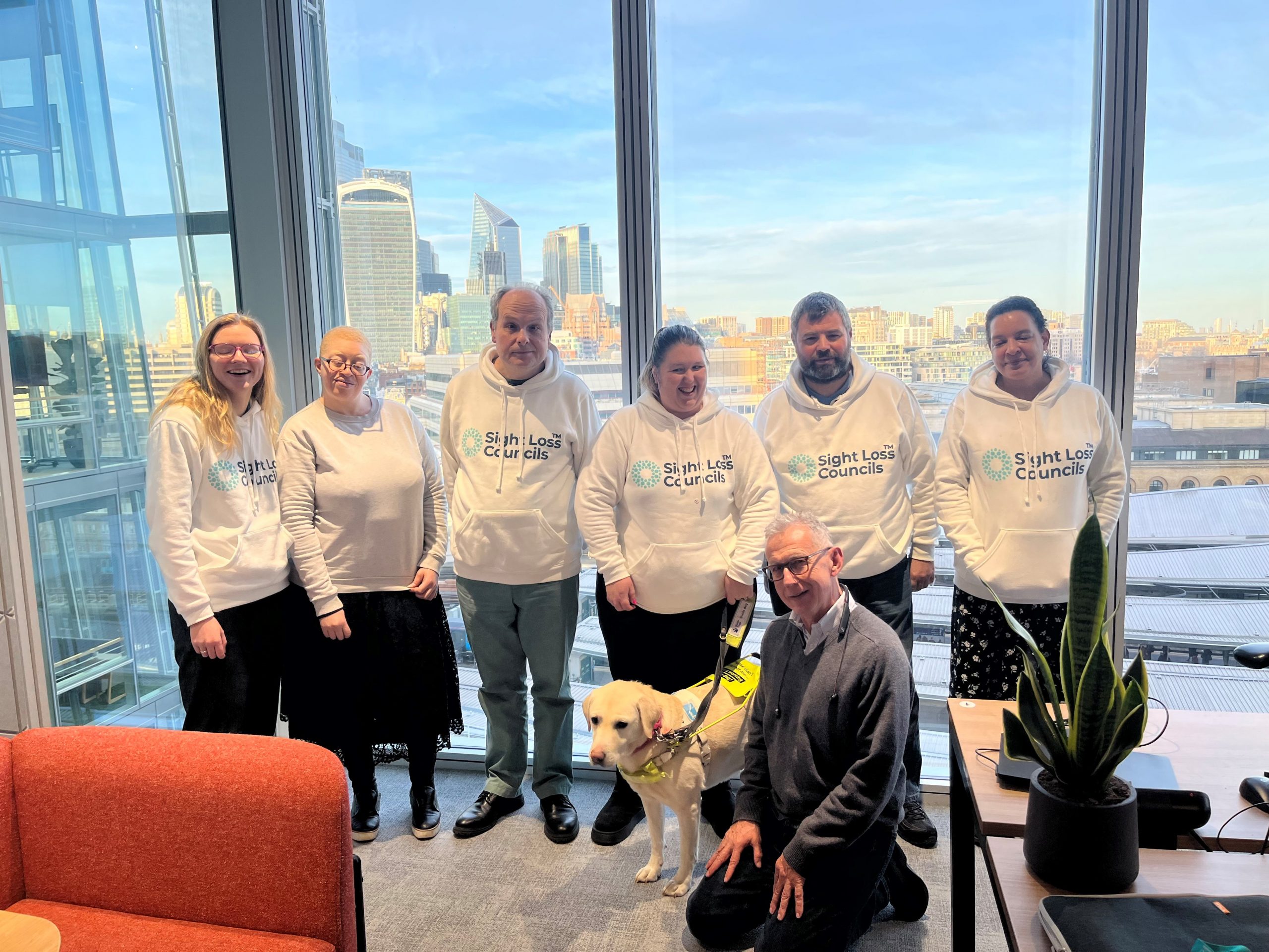 Senior Engagement Manager, Lucy William, pictures with SLC member Emilia, London SLC coordinator Liam, and SLC members Leanne, Steve, and Denise. Peter Richardson who hosted the meeting is knelt on the floor with Guide Dog Dixie.