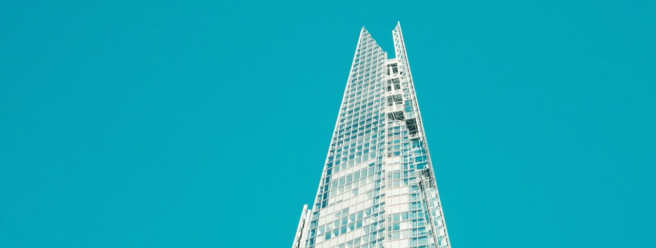 Image looks up at the top third of The Shard, a pyramid shaped, 72 storey, sky scraper, which is based in Southwark, London. The building is glass, and is shown against bright blue sky.