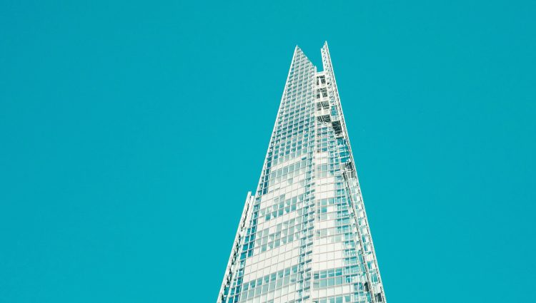 Image looks up at the top third of The Shard, a pyramid shaped, 72 storey, sky scraper, which is based in Southwark, London. The building is glass, and is shown against bright blue sky.