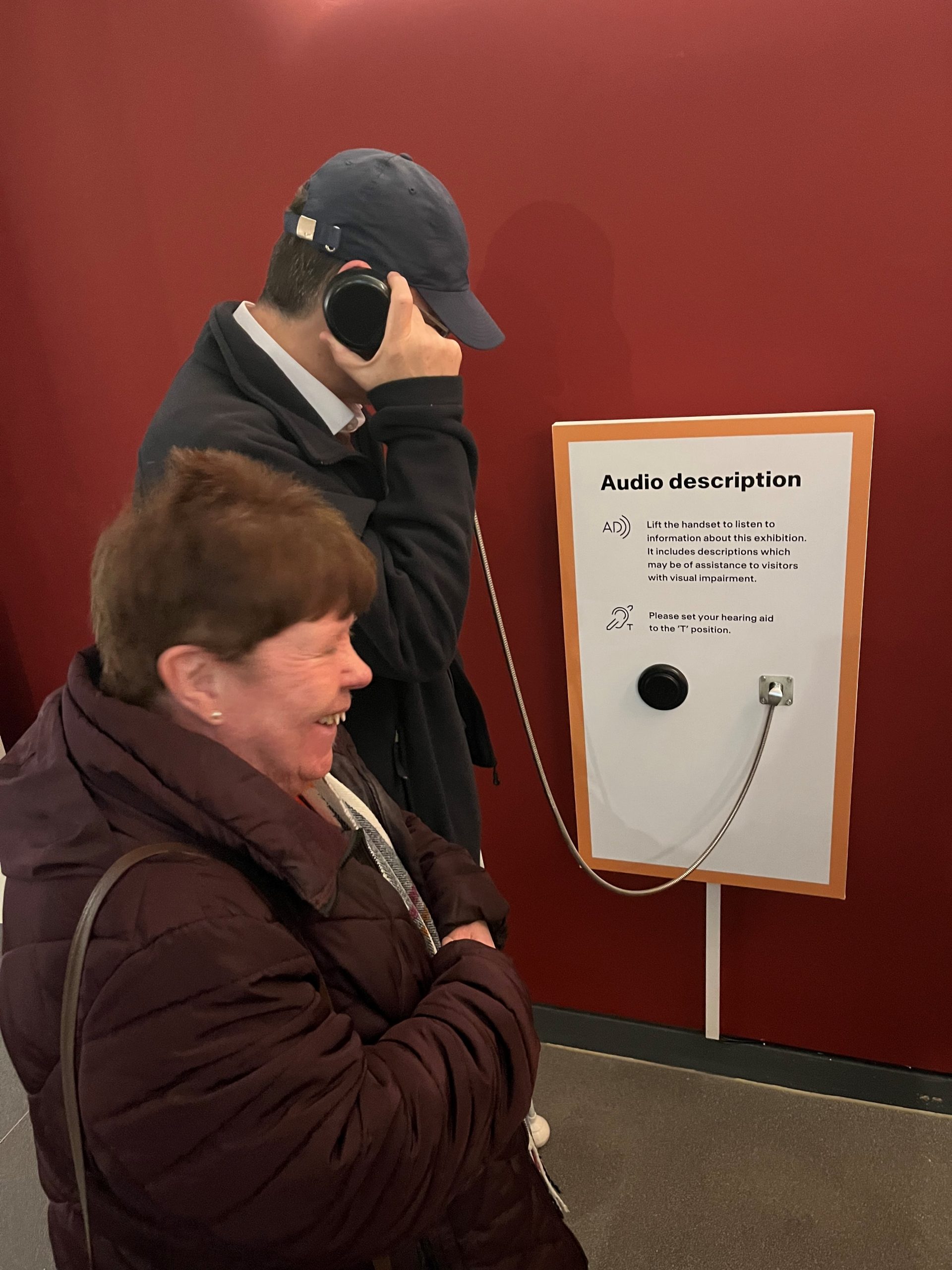 Two delegates pictured using audio description during the Happiness tour. A gentleman is holding a headphone up to his ear whilst a female attendee looks on, smiling.