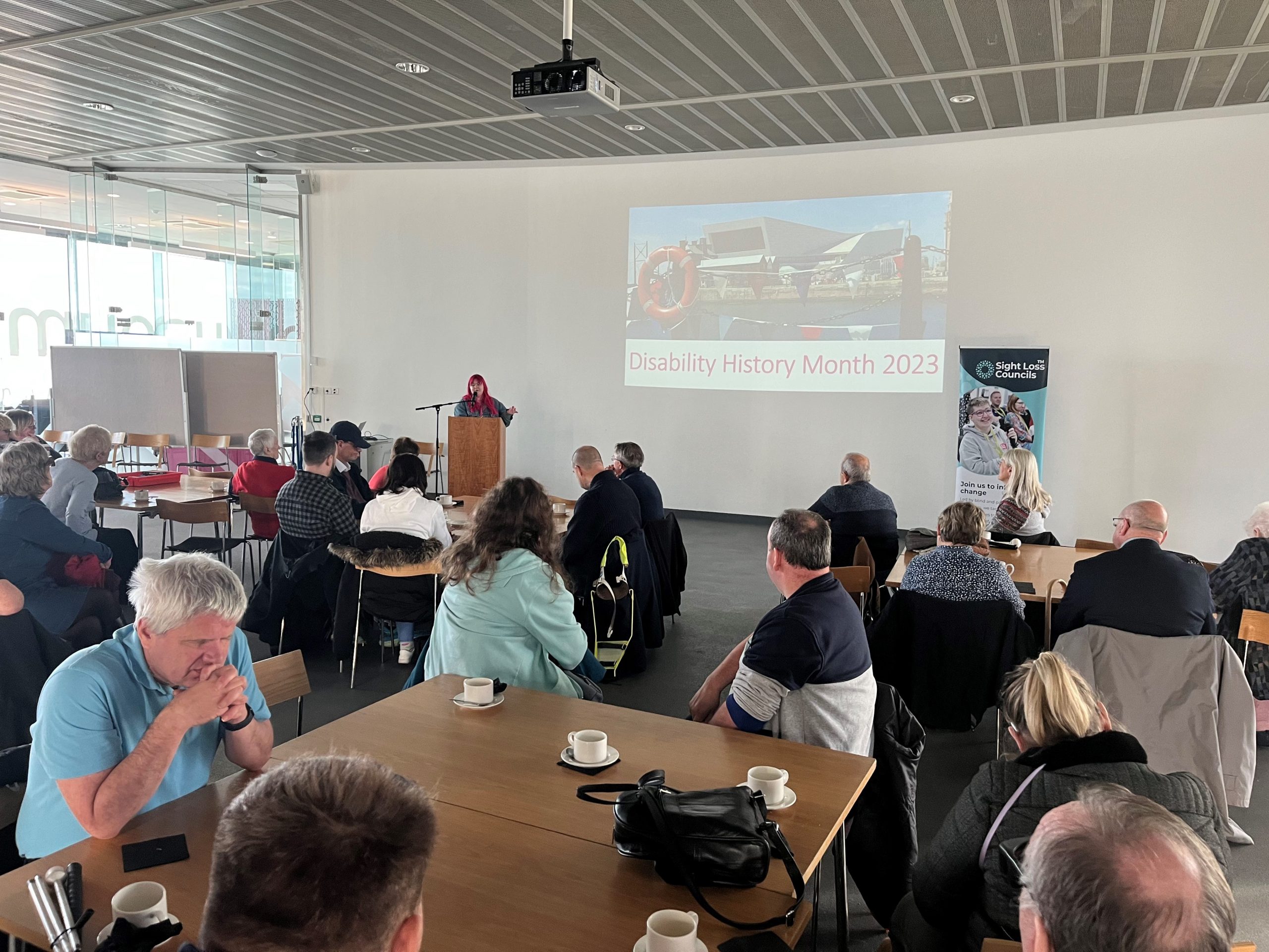 Blind and partially sighted delegates are sat around tables listening to a key speaker at the event. A presentation slide says ‘Disability Awareness Month’ 2023 and there is a Sight Loss Council banner in the background.