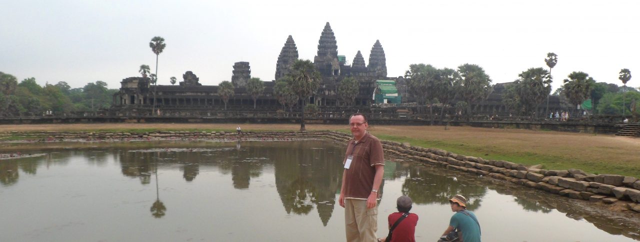 Michael Parkinson, Lancashire SLC member, stood at Angkor Wat in Cambodia. Michael is standing by a stretch of water, with Angkor Wat in the distance.