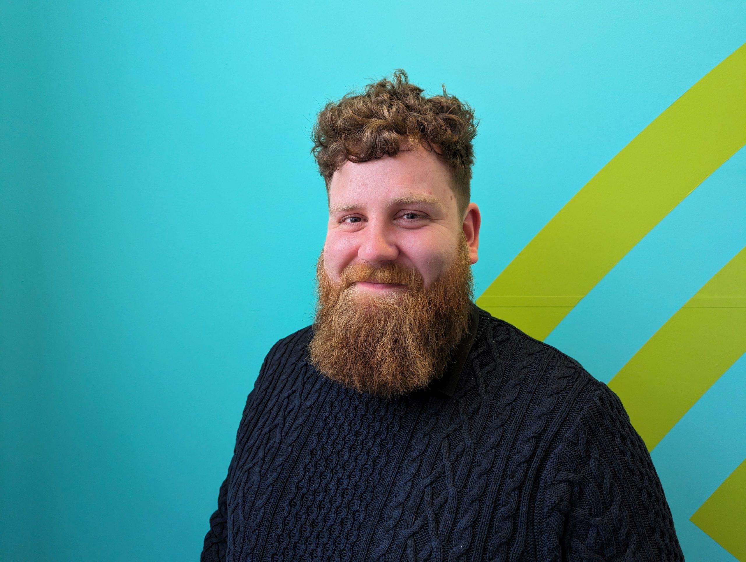 Headshot of Callum Lancashire, Engagement Manager for Scotland. Callum is standing again a turquoise blue background with three yellow strips in the right hand corner. He is wearing a chunky, navy jumper and is turning towards the camera, smiling.